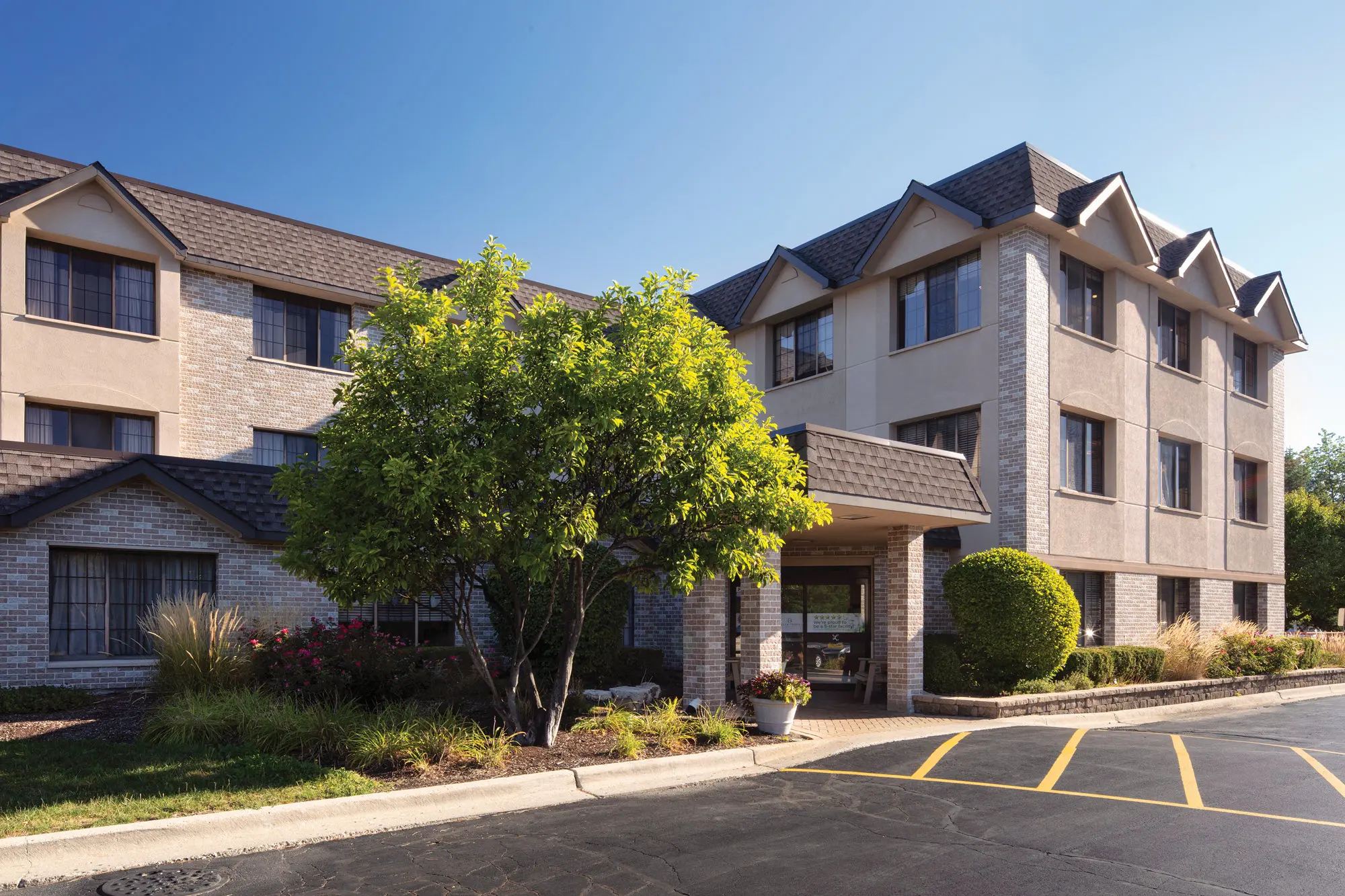 The exterior of a multi-story residential building with a brick and stucco facade, surrounded by landscaped bushes and trees, with a paved parking lot in front and a clear blue sky overhead.