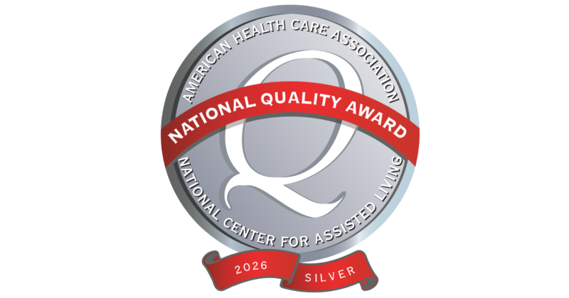 A circular badge with a silver background and a red ribbon banner. The badge reads 