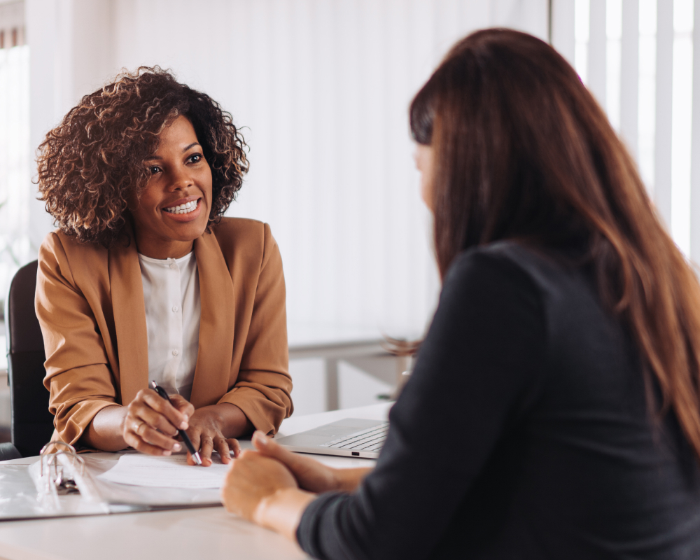 Two women are sitting at a table in a bright office, engaged in a conversation. One woman has curly hair and is smiling while holding a pen, and the other has straight, reddish-brown hair and is listening attentively.