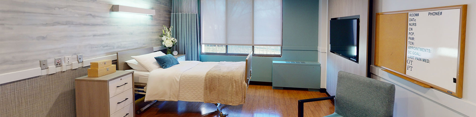 A hospital room with a hospital bed, a bedside table with boxes and flowers, a window with blinds, a wall-mounted TV, and a whiteboard with medical notes.