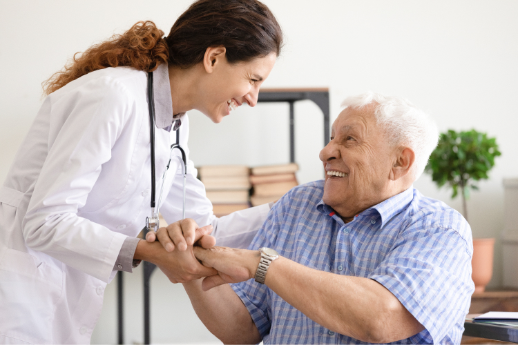 A female healthcare worker smiling and holding hands with an elderly man who is sitting and smiling back.
