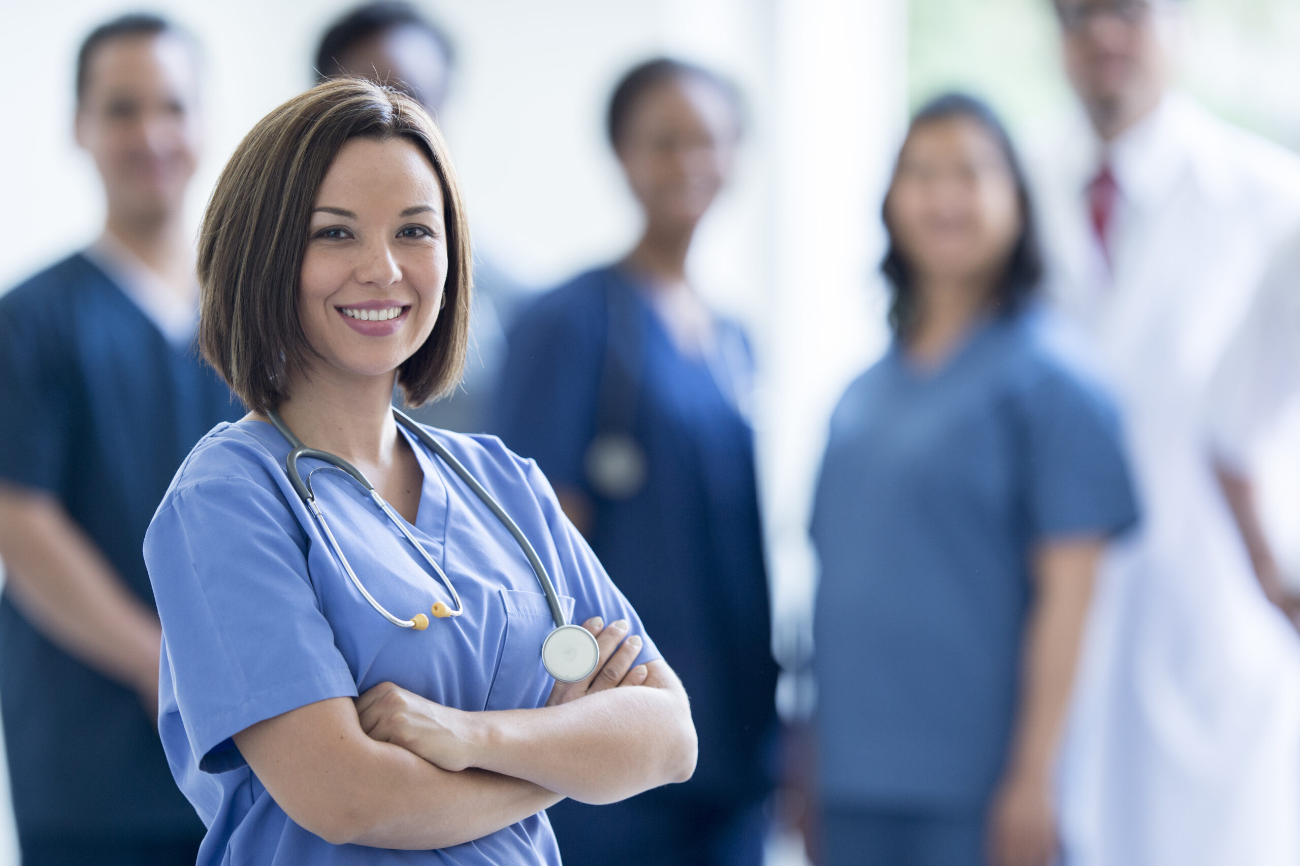 A group of healthcare professionals, including a smiling female nurse in the foreground with arms crossed and a stethoscope around her neck, standing confidently with colleagues in the background.