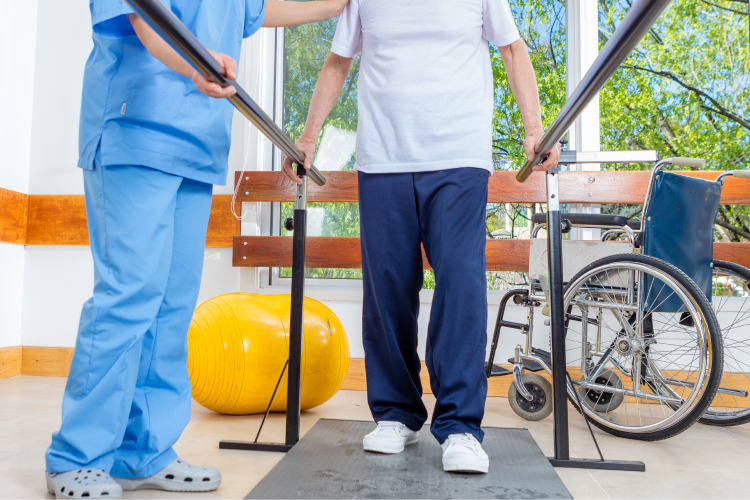 A person using parallel bars for rehabilitation, assisted by a healthcare worker in scrubs, with a wheelchair and exercise ball nearby in a sunlit therapy room.