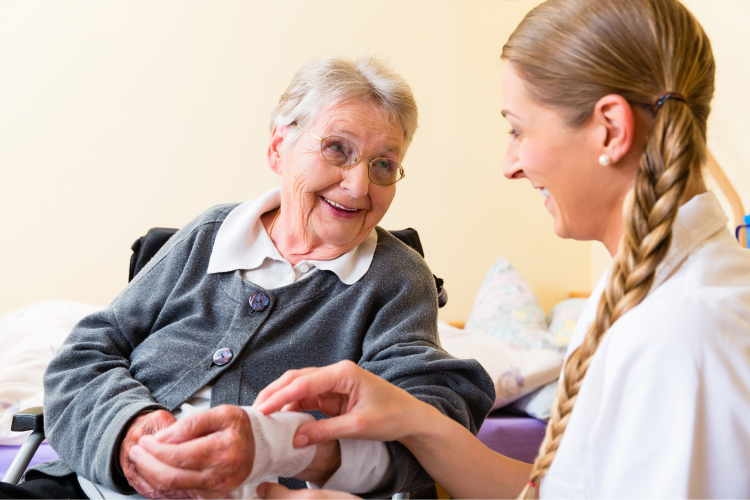 A smiling elderly woman with glasses sits in a wheelchair, holding hands with a young female caregiver who is smiling and gently touching her hand, in a warm, caring interaction.