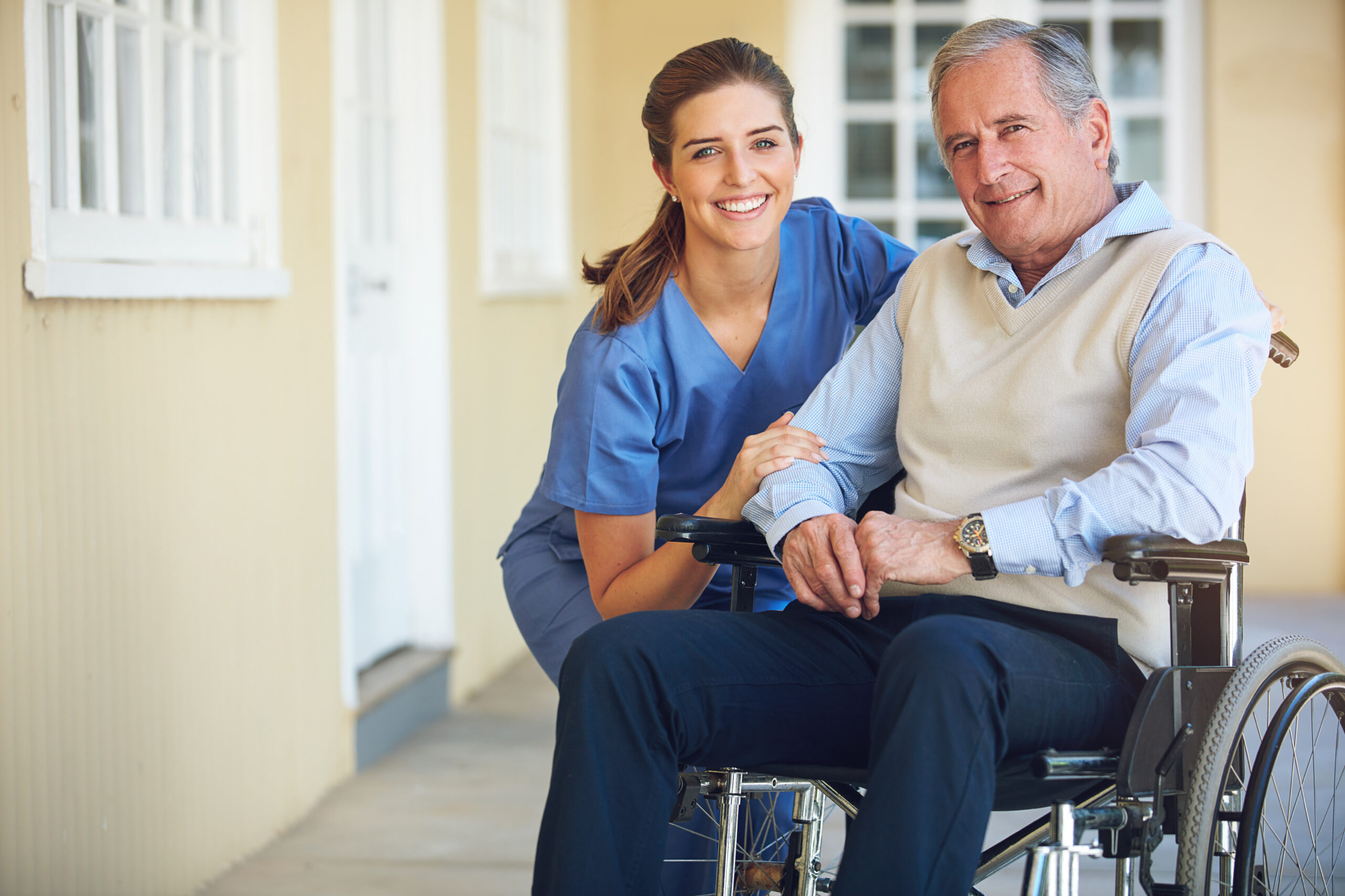 A smiling young female healthcare worker in blue scrubs supports an elderly man in a wheelchair, both sitting in a brightly lit hallway with yellow walls and white window frames.