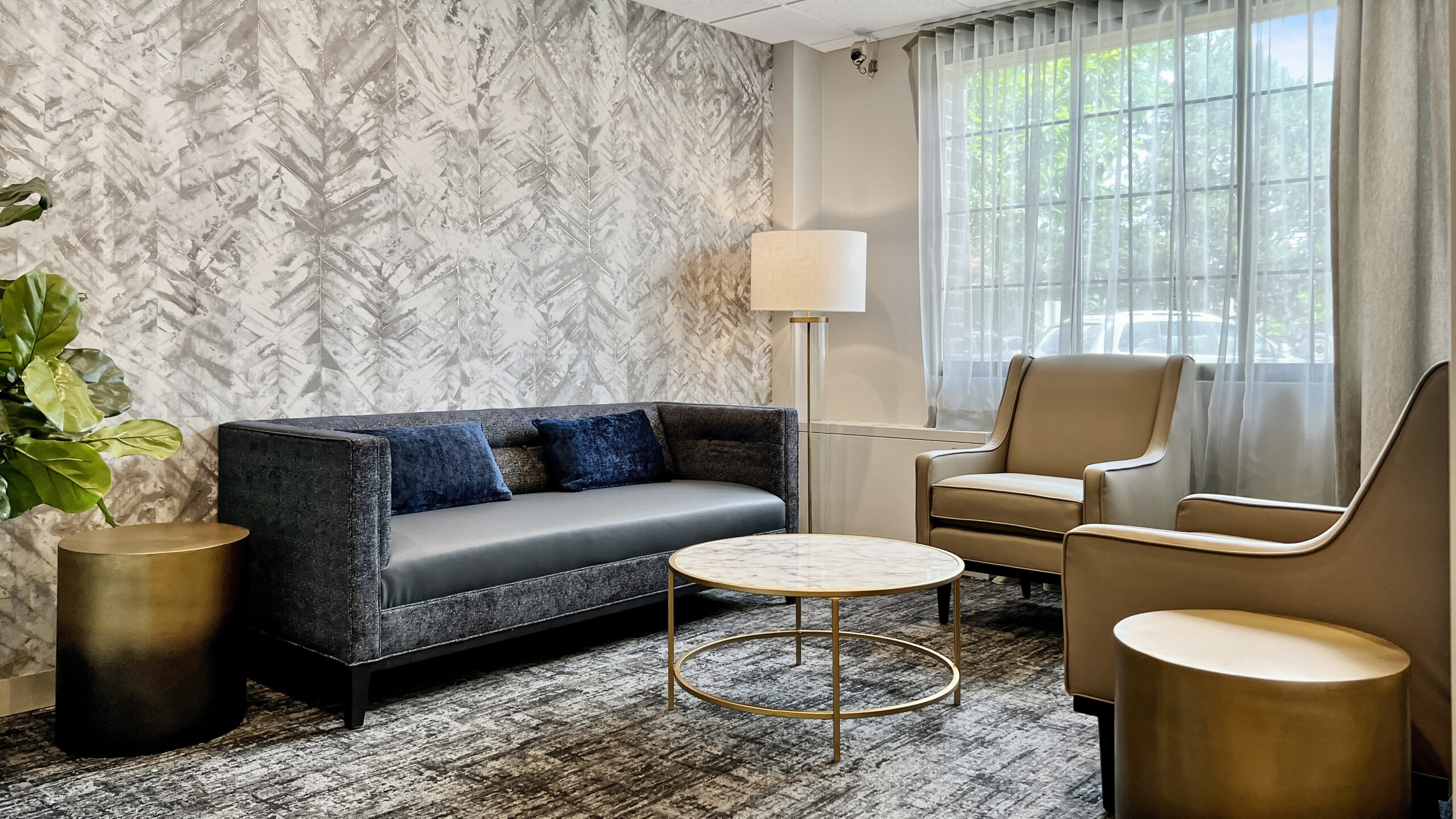 A cozy hotel sitting area with a dark gray sofa, two beige armchairs, a round marble-topped coffee table with a gold frame, a tall white floor lamp, a large window with sheer white curtains, and patterned wallpaper featuring a leaf design. There is also a green potted plant on the left side.