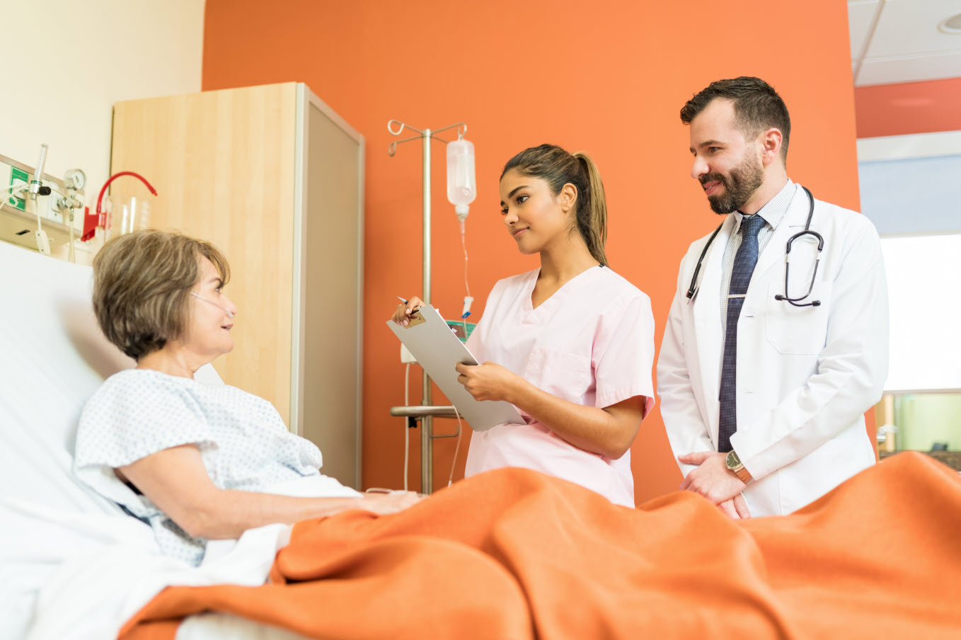 A female patient lying in a hospital bed, talking to a male doctor and a female nurse who are standing beside her. The nurse is holding a clipboard and taking notes. The background features medical equipment and a bright orange wall.