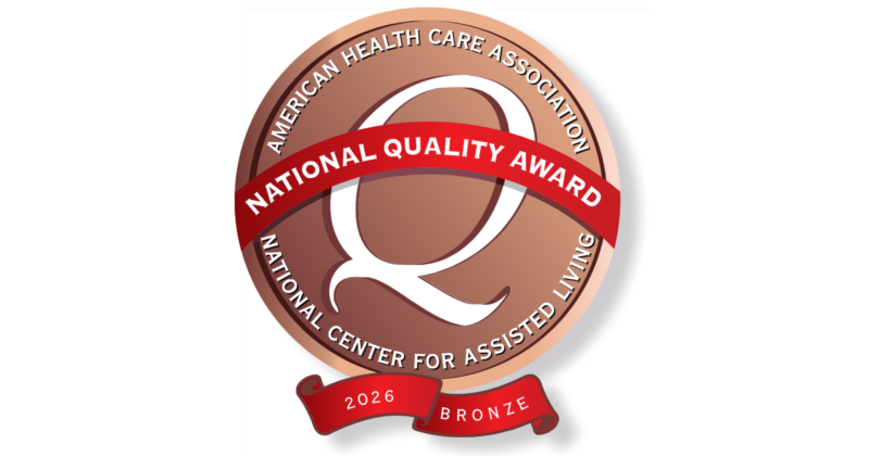 A circular award badge with a bronze background and a red ribbon across the center. The ribbon reads "NATIONAL QUALITY AWARD" in white letters. The outer edge of the badge has the text "AMERICAN HEALTH CARE ASSOCIATION" at the top and "NATIONAL CENTER FOR ASSISTED LIVING" at the bottom. Inside the badge, a large white letter "Q" is displayed. A smaller red banner at the bottom of the badge reads "2026 BRONZE".