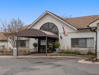 A single-story building with a white exterior and a large arched window above the entrance. The entrance has a black awning and glass doors, with small trees and shrubs on either side. A parking lot is in the foreground, and the sky is clear and blue.
