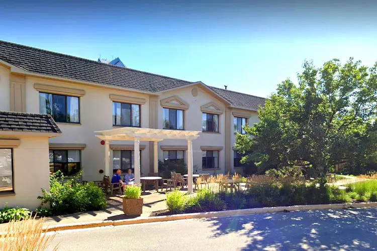 A two-story beige building with multiple windows, surrounded by greenery and trees. There is an outdoor seating area with tables, chairs, and a white pergola, with a few people sitting and chatting.