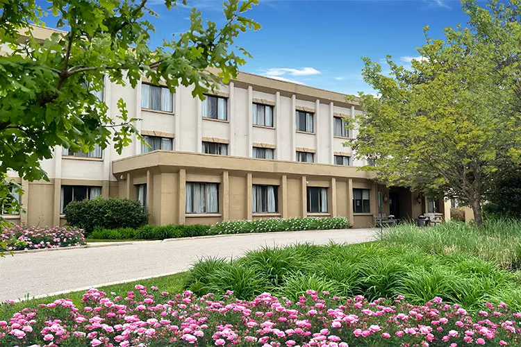 A multi-story beige hotel building with several windows, surrounded by green trees and landscaped gardens with pink flowers and lush grass, under a bright blue sky with a few clouds.