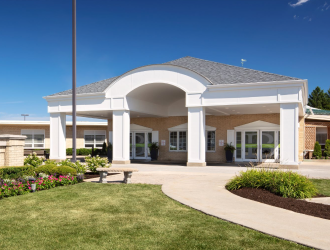 The image shows the exterior of a building with a large, white, arched entrance supported by columns. There is a well-maintained lawn with flower beds and a paved walkway leading to the entrance. The sky is clear and blue.