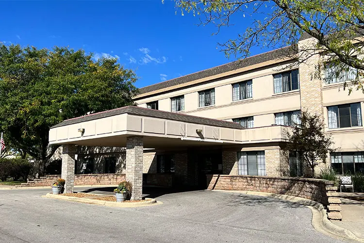 A multi-story beige hotel building with dark window frames, featuring a covered entrance supported by brick columns, surrounded by trees and potted plants, under a bright blue sky.