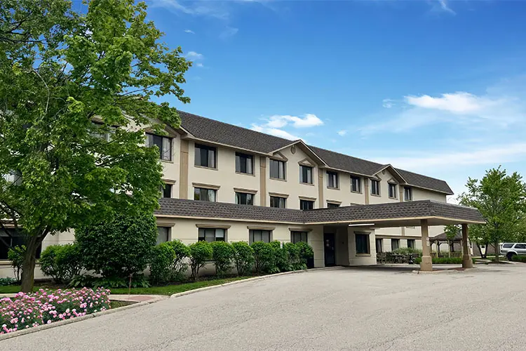 A four-story beige hotel building with dark brown roof, surrounded by green trees and bushes, with a covered entrance and a parking area in front under a blue sky with some clouds.